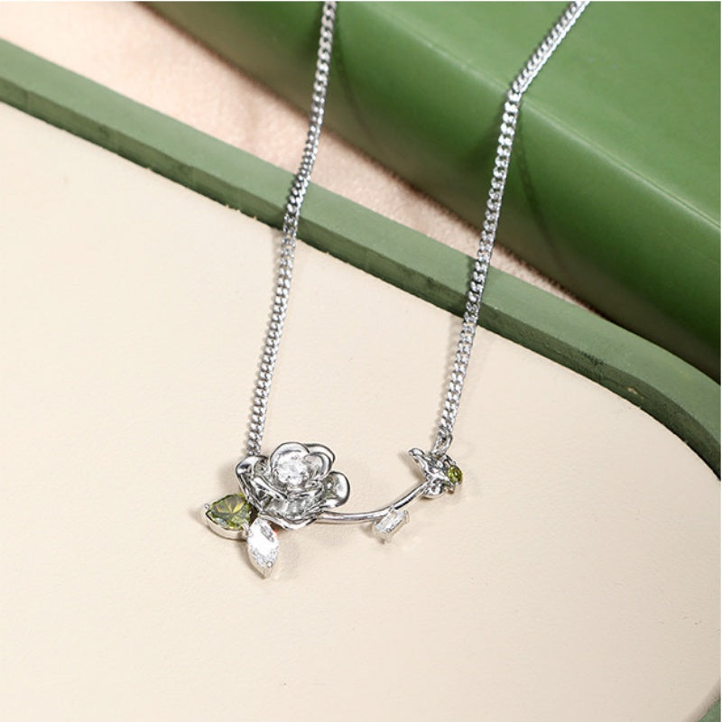 Close-up of a silver-tone sculpted rose necklace with green leaf details and a small dangling charm.