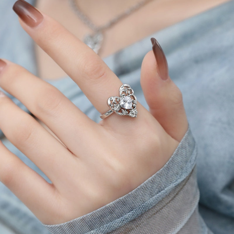 Close-up of a silver-tone ornate openwork heart ring with a clear central stone, worn on a hand.