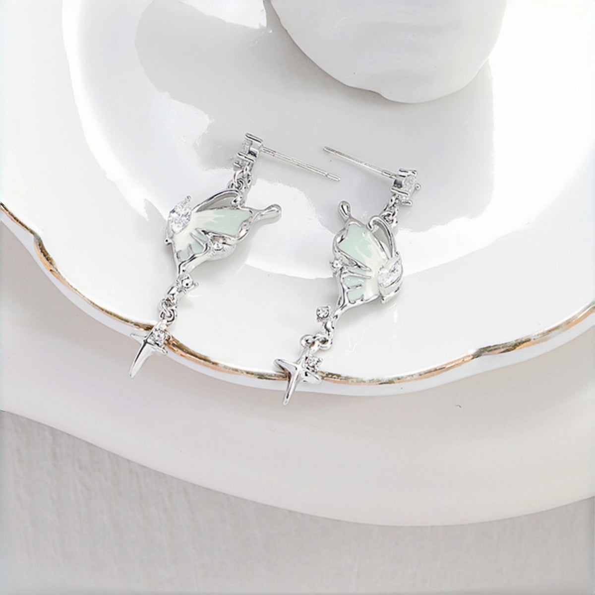 Fairy iridescent butterfly drop earrings close-up, pair of silver butterfly charms with mint enamel wings and clear crystal accents, on a white plate