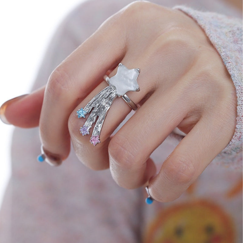Close-up of a white star ring with silver-tone shooting-star trails tipped with pale blue and pink details, worn on a hand.