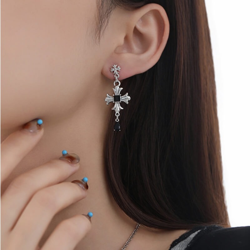 Close-up of a silver-tone cross drop earring with a black square centre and a dark dangling bead.