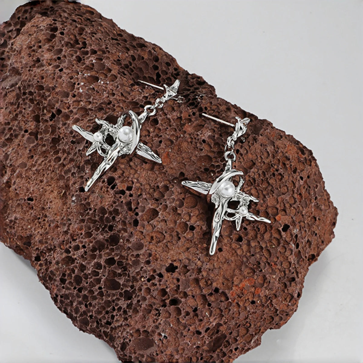 Grunge star cross drop earrings close-up, pair of silver four-point star and cross charms with pearl accents, on a textured brown rock