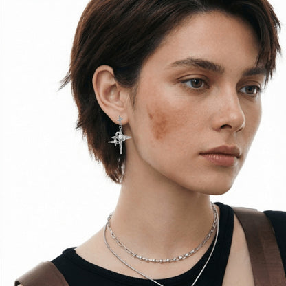 Close-up of a silver-tone star-cross drop earring with long pointed arms, worn by a model.