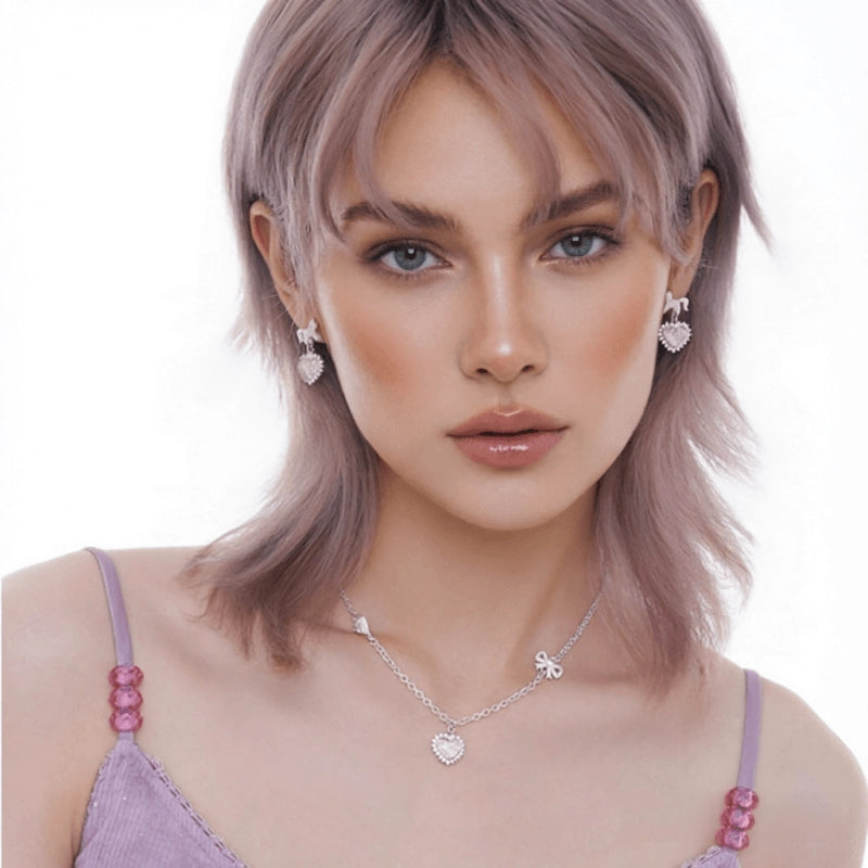 Model wearing silver-tone heart drop earrings and a matching heart pendant necklace with small bow details.
