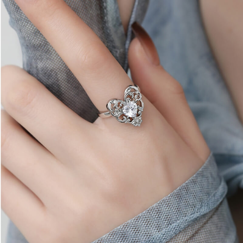 Close-up of a silver-tone ring with an ornate openwork heart and a clear central stone, worn on a hand.