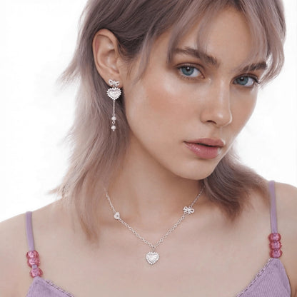 Model wearing a pale pink heart pendant necklace and a bow-and-heart earring with a long pearl-like drop.