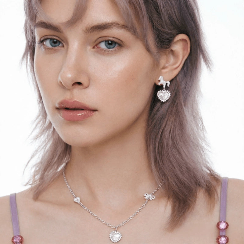 Model wearing a pale pink heart pendant necklace and a short silver-tone drop earring with a heart charm.