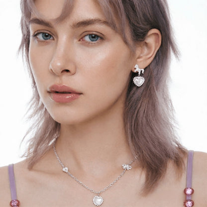 Model wearing a pale pink heart pendant necklace and a short silver-tone drop earring with a heart charm.