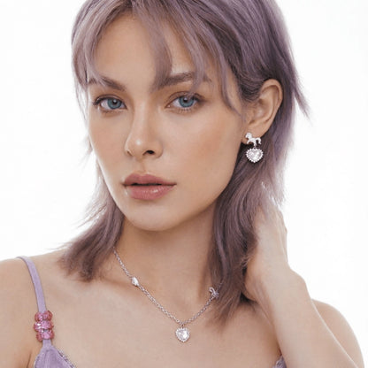 Model wearing a pale pink heart pendant necklace and matching unicorn drop earring in silver-tone settings.