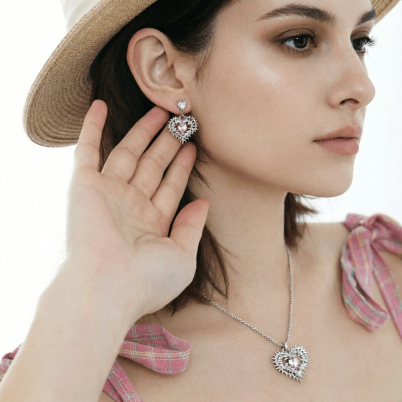 Model wearing a silver-tone thorn-framed heart earring and matching necklace with pale pink centres.