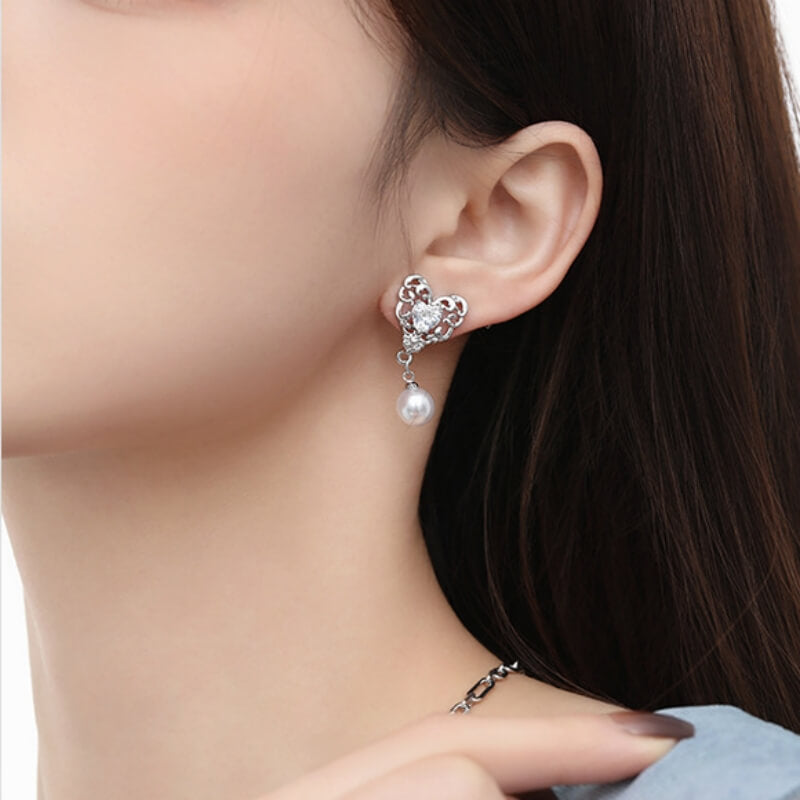 Close-up of a silver-tone ornate heart earring with a clear central stone and a pearl-like drop, worn by a model.