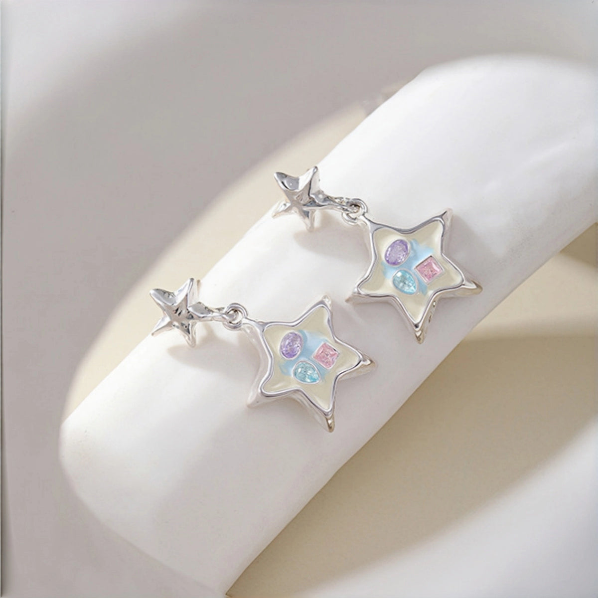 Enamel star drop earrings close-up, pair of silver star studs with enamel star charms featuring pastel crystals, resting on a display stand