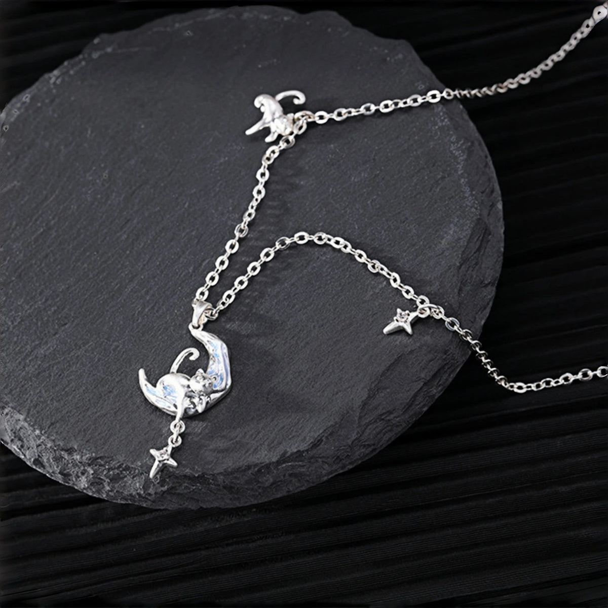 Moon and cat necklace close-up, silver crescent moon pendant with a curled cat charm and dangling star, on a slate surface