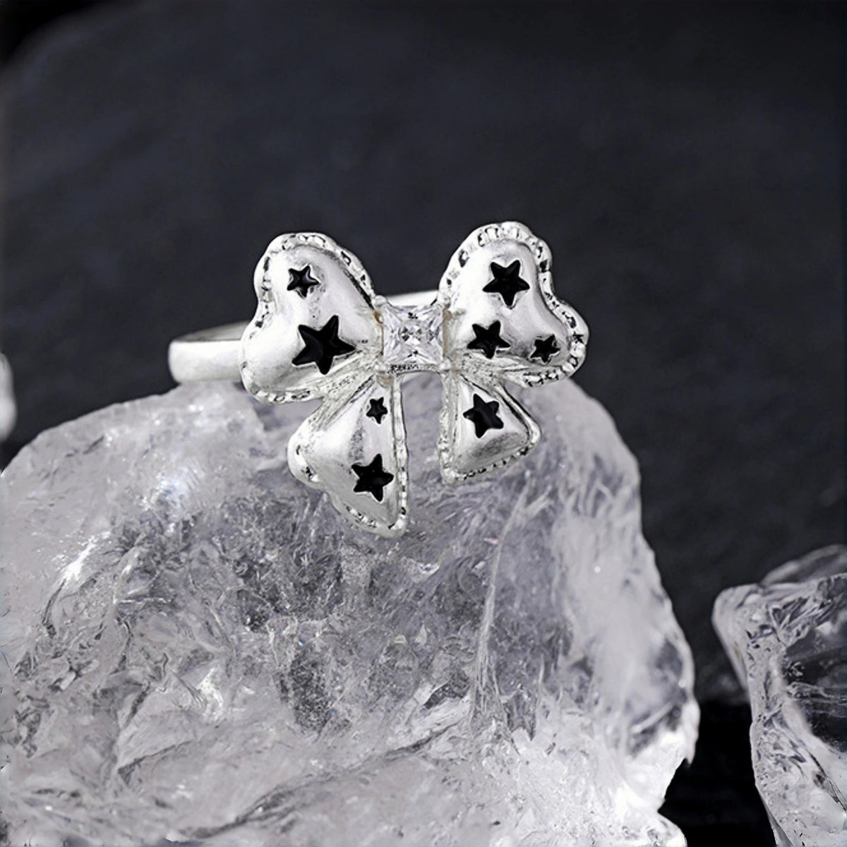 Silver bow ring close-up, open band with a bow charm featuring black star cutouts, resting on a clear crystal rock