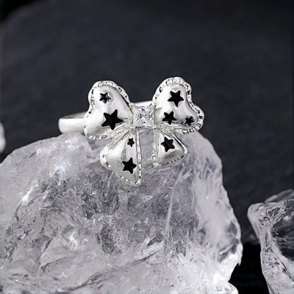 Silver bow ring close-up, open band with a bow charm featuring black star cutouts, resting on a clear crystal rock
