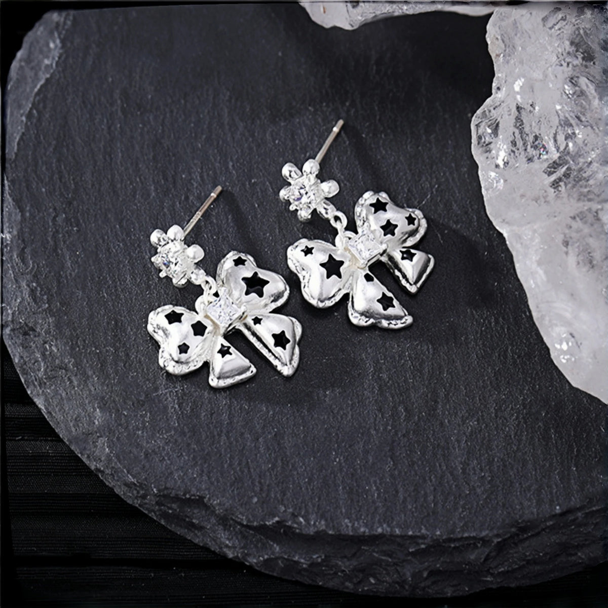 Silver bow drop earrings close-up, pair of bow charms with black star cutouts and small crystal stud tops, on a slate surface beside crystal rocks
