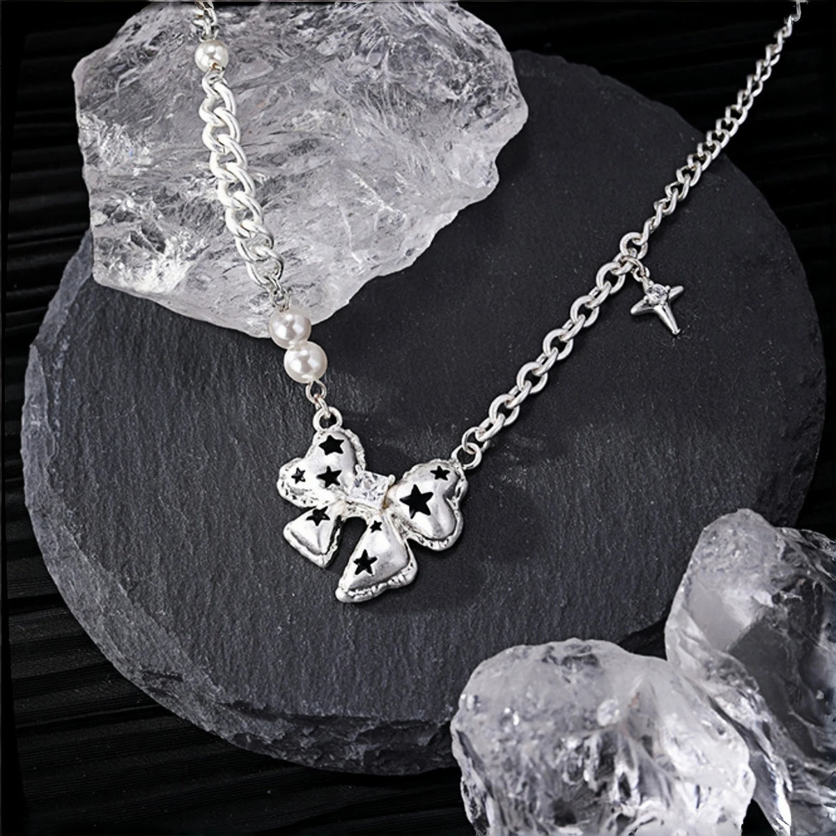 Silver bow pendant necklace close-up, bow charm with black star cutouts on a curb chain with pearl beads and a dangling star charm, on a slate surface with crystal rocks