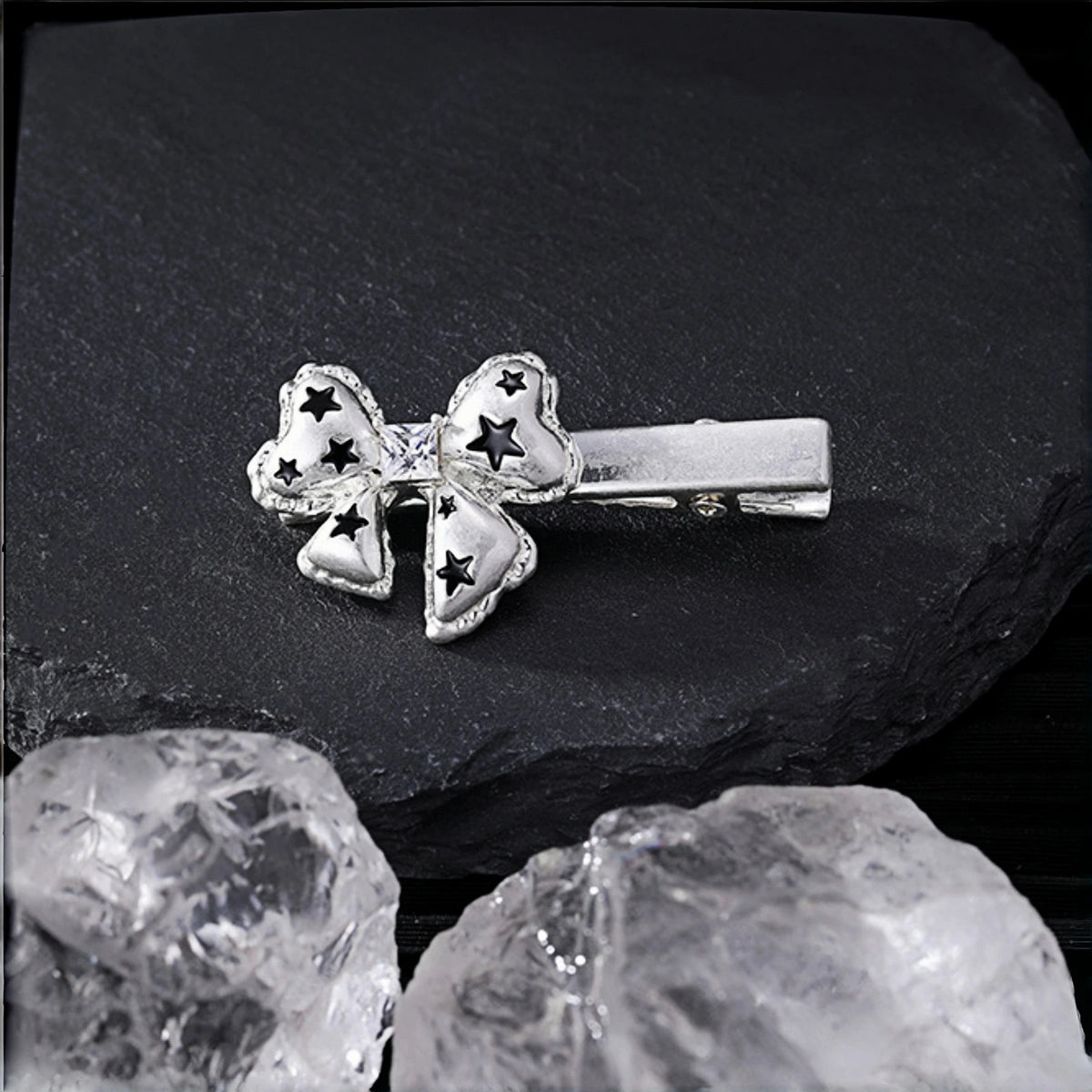 Silver bow hair clip close-up, alligator clip topped with a bow charm featuring black star cutouts, on a slate surface with crystal rocks