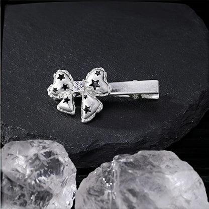 Silver bow hair clip close-up, alligator clip topped with a bow charm featuring black star cutouts, on a slate surface with crystal rocks